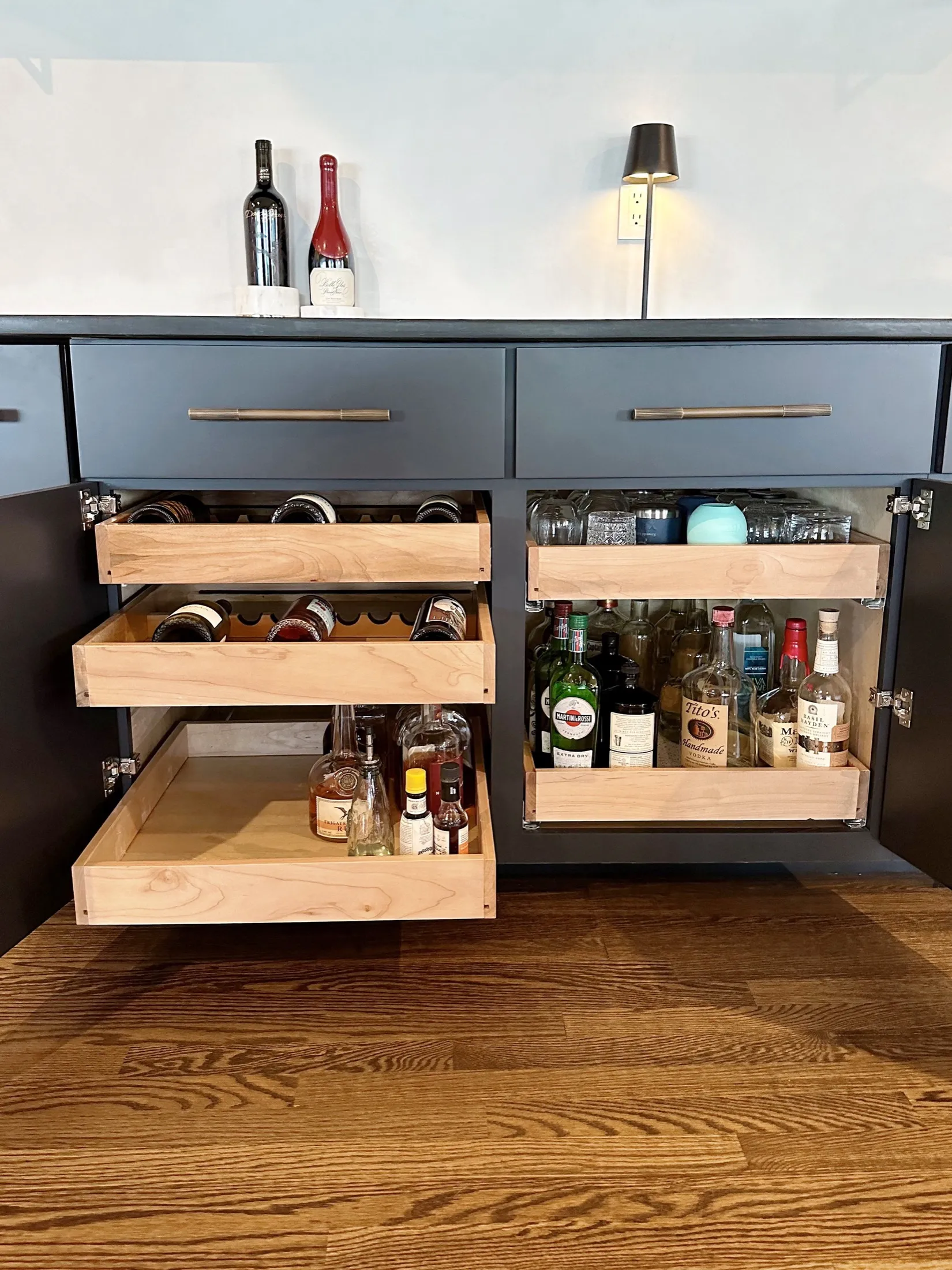 Open black cabinet with beer and liquor bottles, glasses, and wine bottles inside, on a wooden floor, with decorative bottles and a lamp on top.
