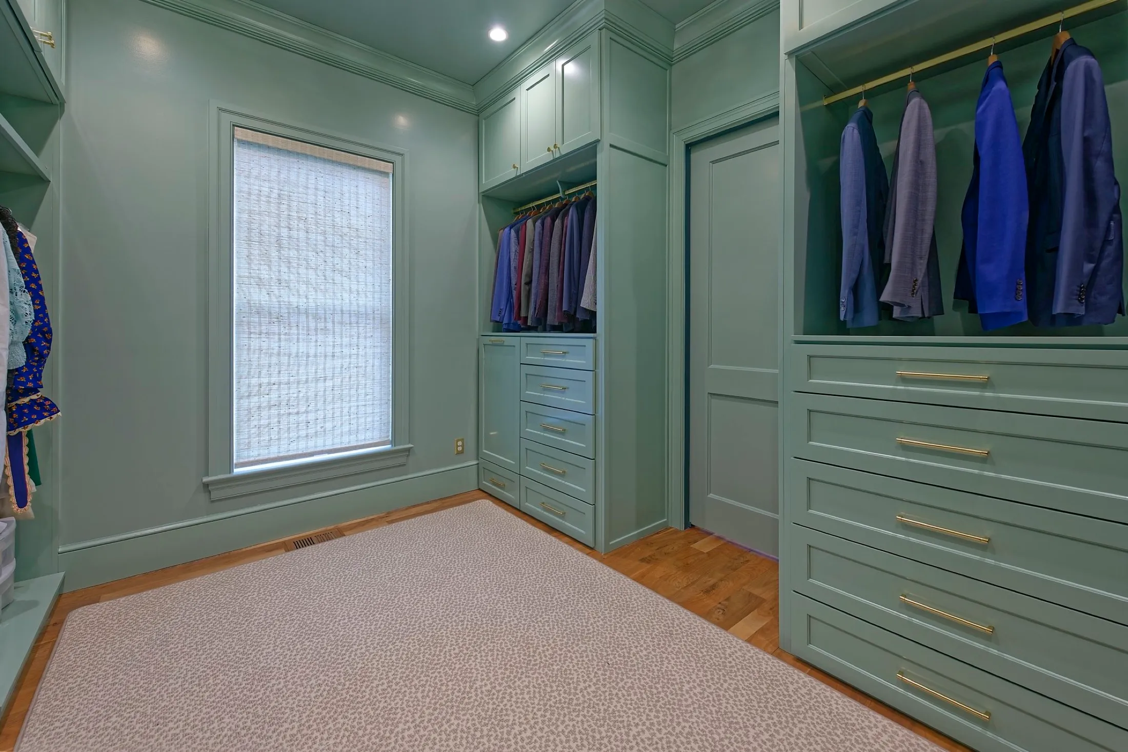 A walk-in closet with green built-in cabinets and drawers, hanging suits, and a window with a woven shade.