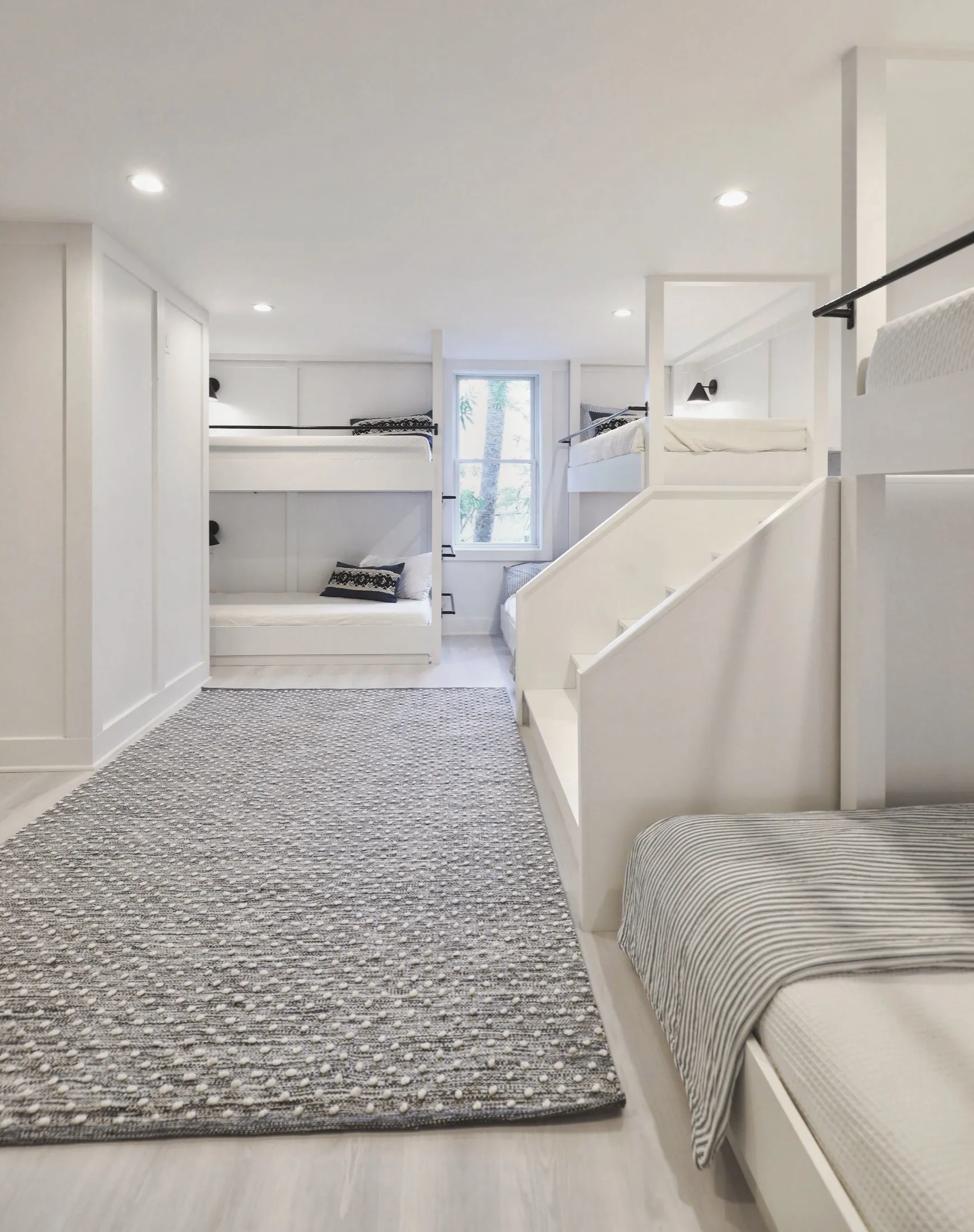 A bedroom with four white bunk beds, a gray and white patterned rug on light wooden floors, a window with plants outside, and a staircase leading to the lower bed.