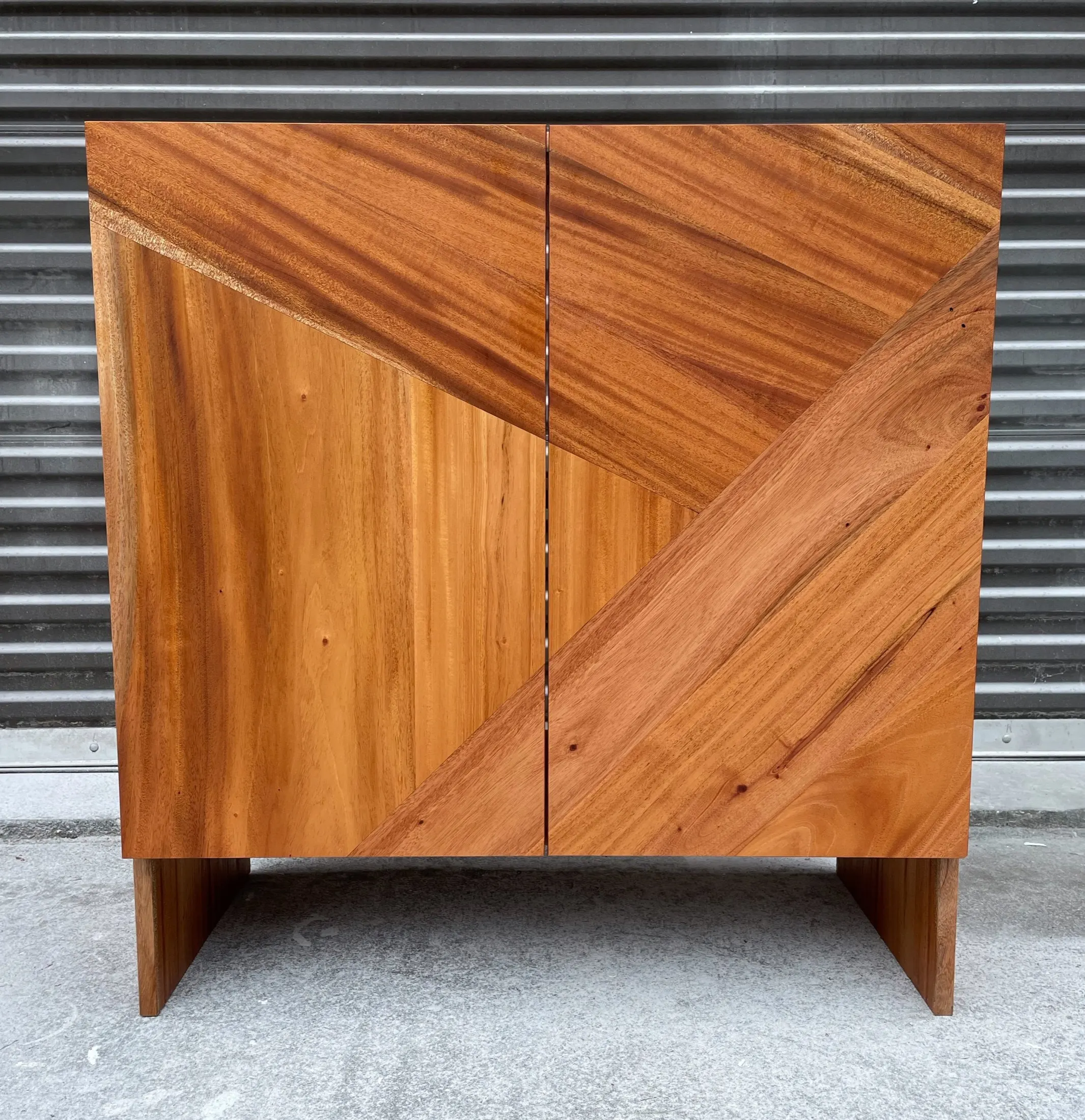 Wooden cabinet with diagonal and vertical wood grain patterns, standing on a concrete floor against a metal wall.