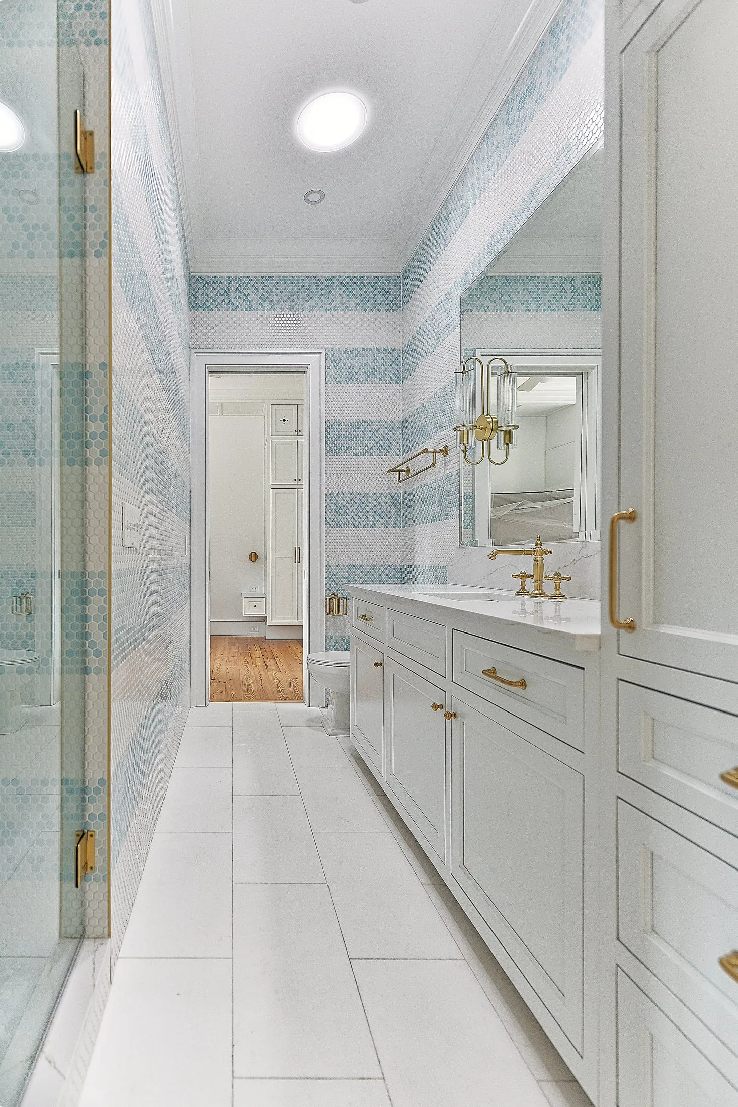 Long bathroom with a white vanity and linen cabinet, glass shower and blue striped tile.