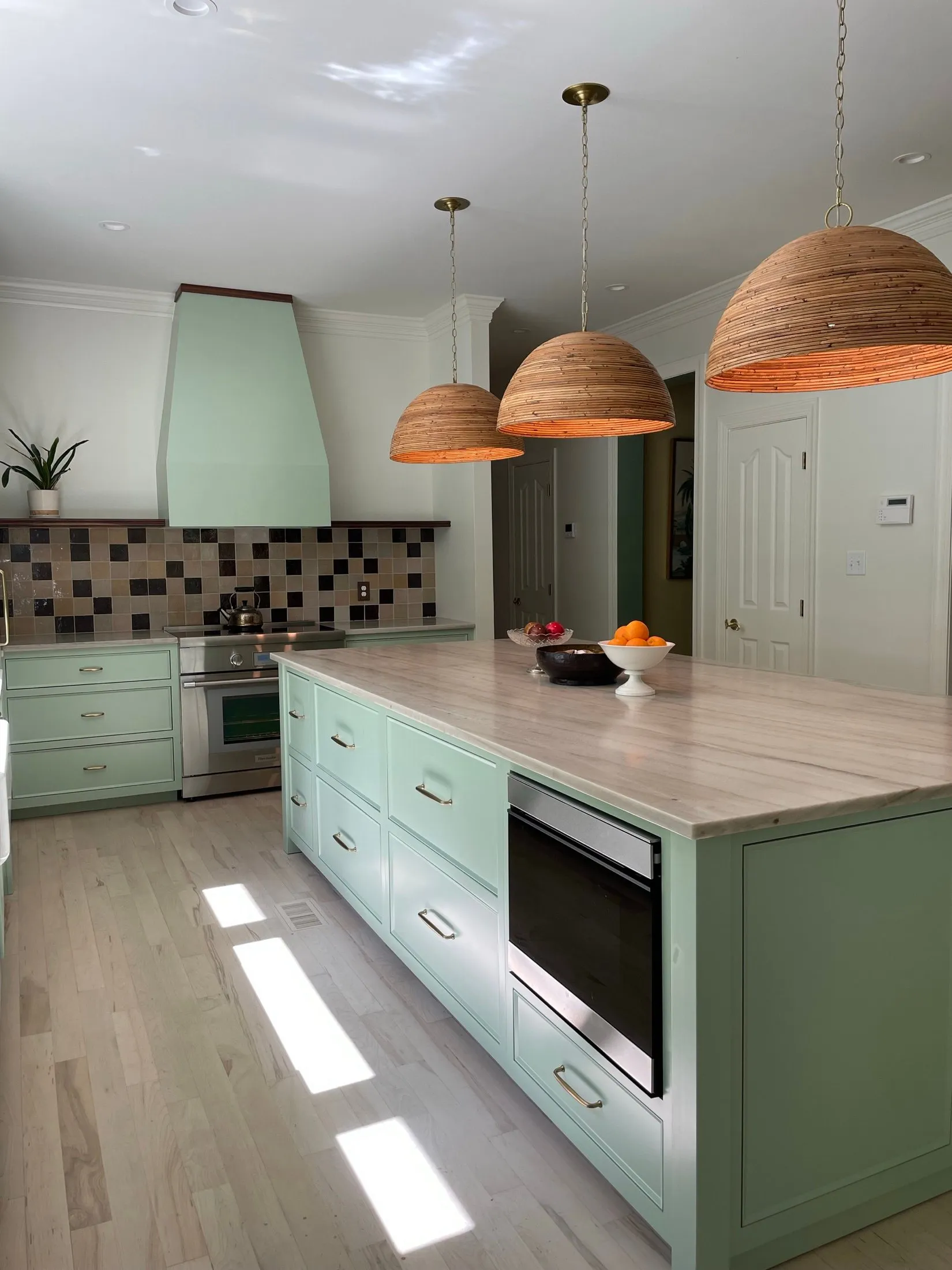 Kitchen with mint green cabinets, wooden countertop, three wooden pendant lights hanging from the ceiling, a bowl of oranges, a stove with an oven, a plant on the counter, and sunlight on the hardwood floor.