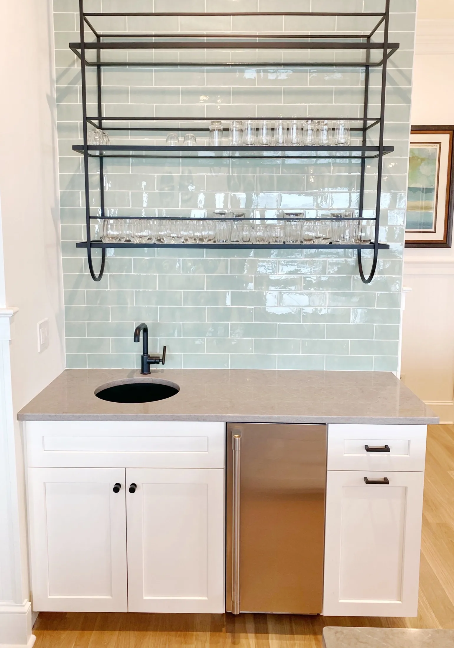 A small bar area with a light green tiled backsplash, white cabinetry, a black faucet, a small sink, and a stainless steel mini fridge. Open black metal shelves above hold drinking glasses.