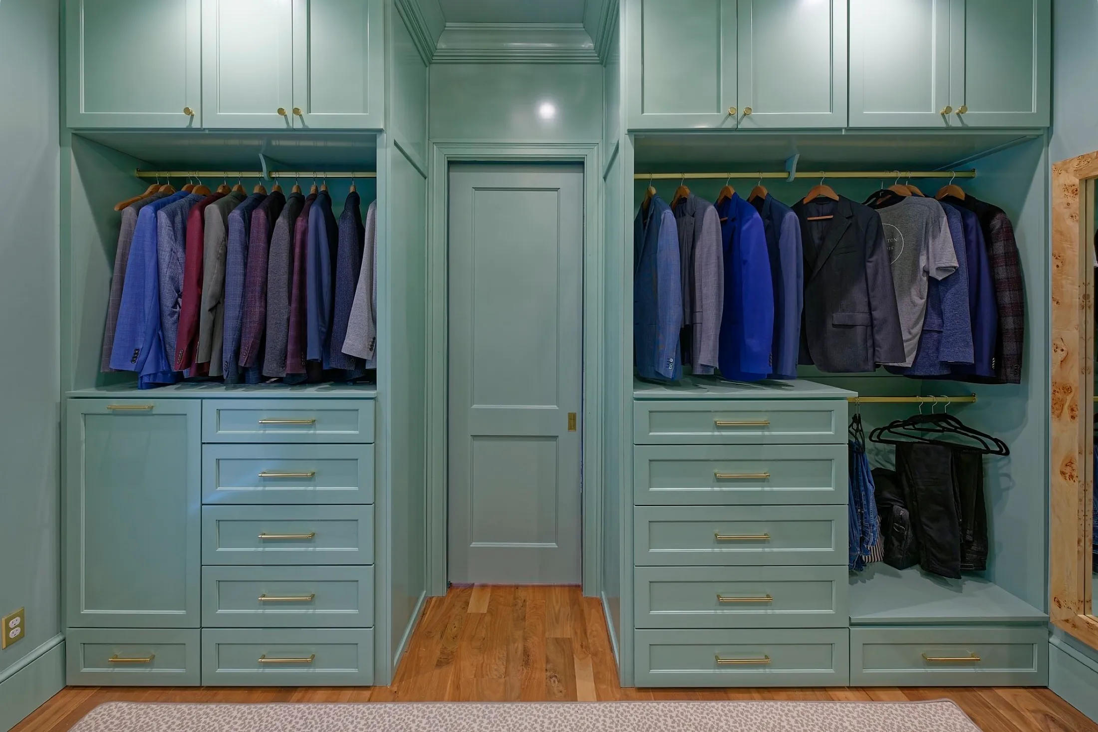 A walk-in closet with mint green built-in cabinets and drawers, filled with hanging suits, blazers, shirts, and pants, on gold hangers, with hardwood flooring and a beige rug.