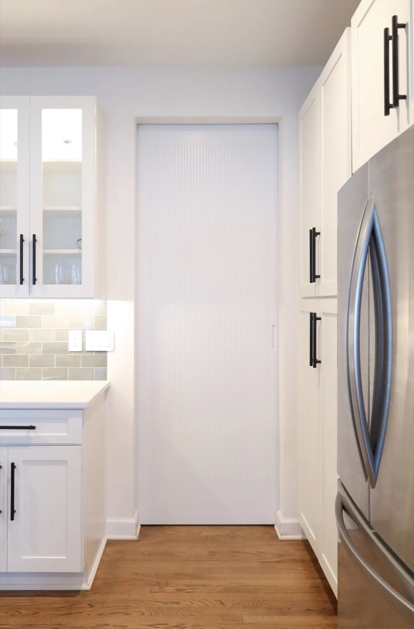 Kitchen with white cabinets, a sliding white door, stainless steel refrigerator, hardwood flooring, and beige tile backsplash.