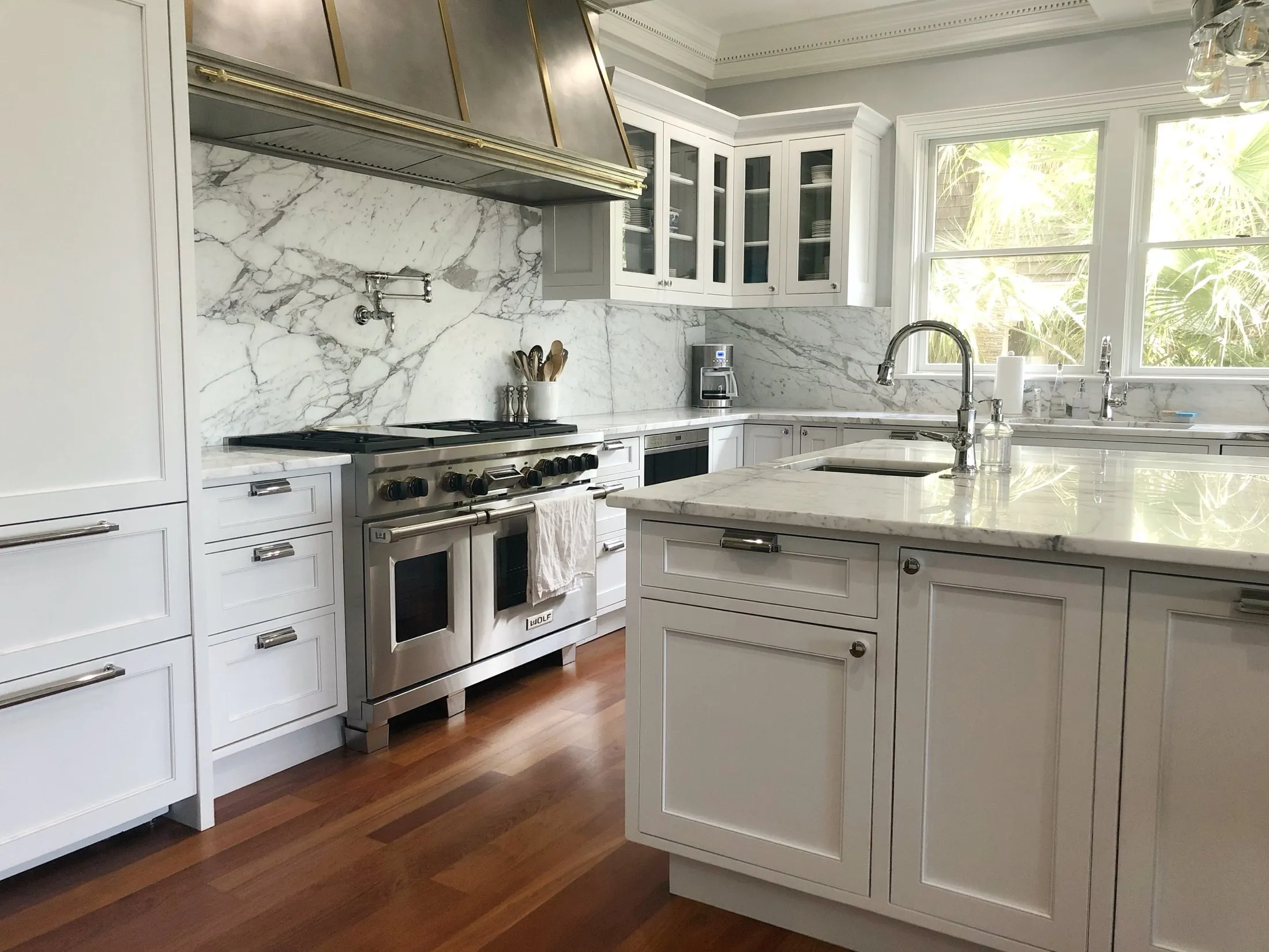 A bright, modern kitchen with white cabinetry, marble countertops, and backsplash. There is a stainless steel stove with oven, a coffee maker, and a large window overlooking greenery. The kitchen island has a sink and soap dispenser, with hanging gla