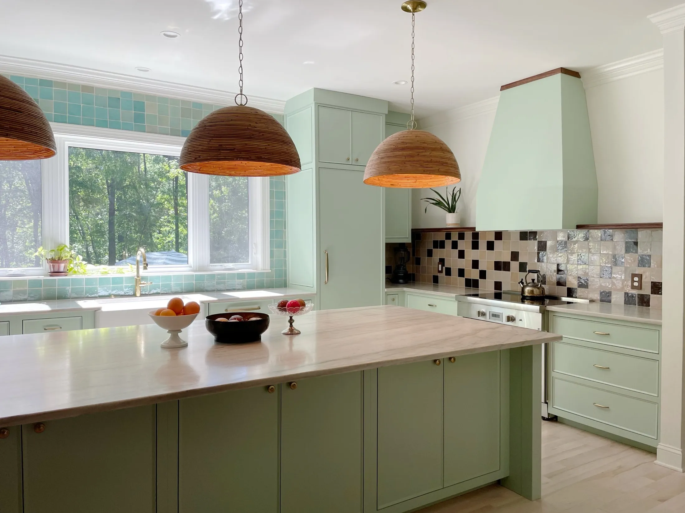 A light green kitchen with a large wooden island, three wicker pendant lights, a window with trees outside, and a tiled backsplash behind the stove.