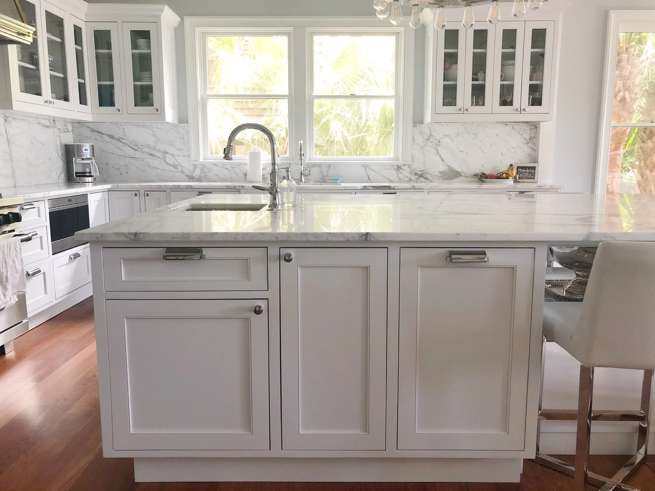 Bright kitchen with white cabinets, marble countertops, a kitchen island, and large windows with greenery outside