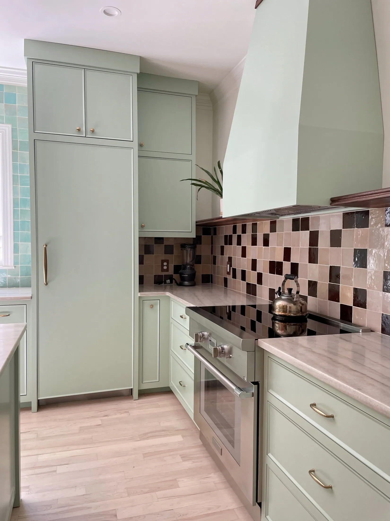 A kitchen with light green cabinets, a multicolored tiled backsplash, a stove with a metallic kettle on top, a potted plant, and a blender on the counter