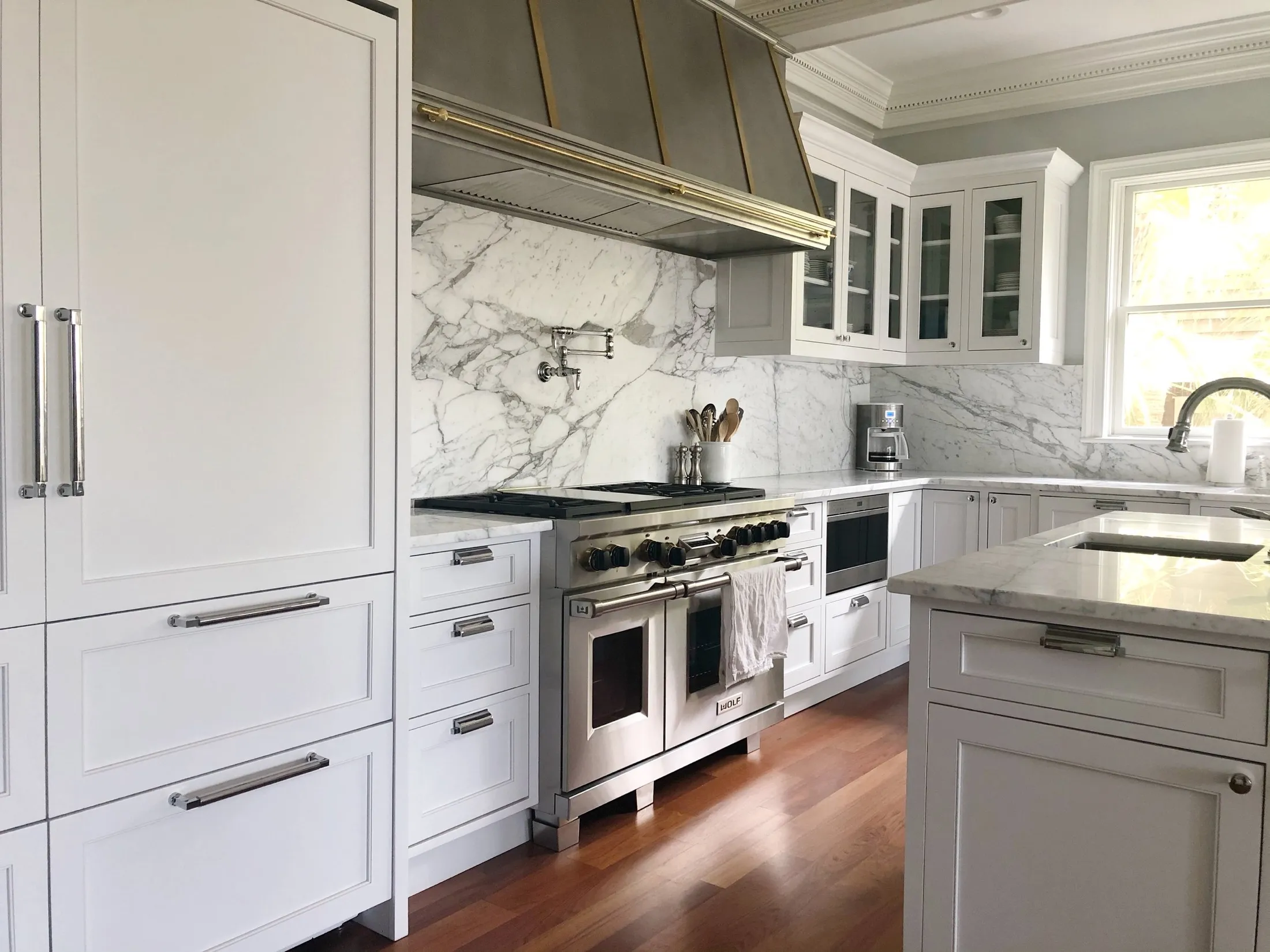 Bright, modern kitchen with white cabinets, marble countertops and backsplash, stainless steel appliances including oven and microwave, a coffee maker, and a large window letting in natural light.