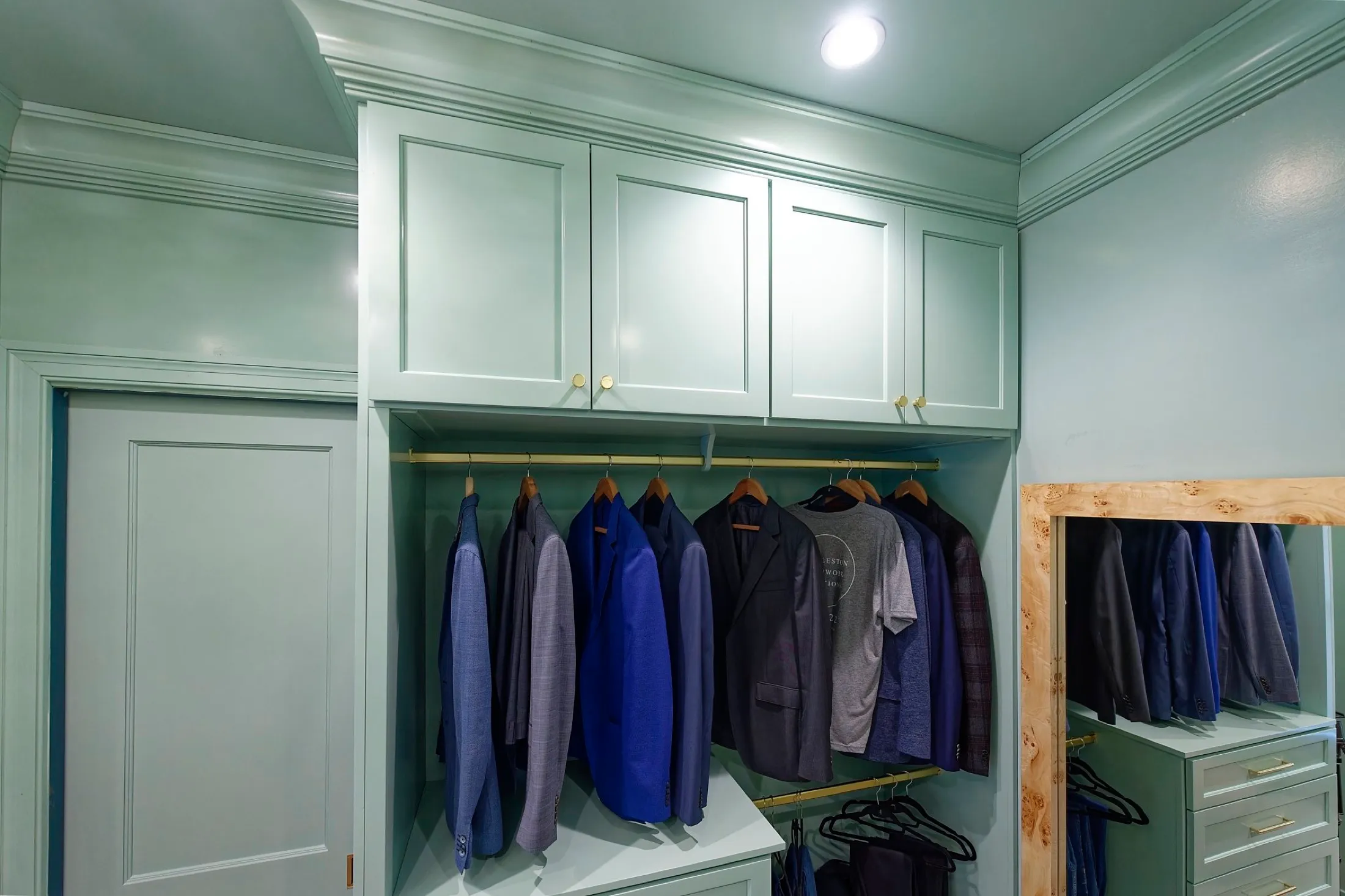 Closet with hanging suits, shirts, and jackets, white cabinets above, a small dresser to the right, and wooden framing around the dresser area.
