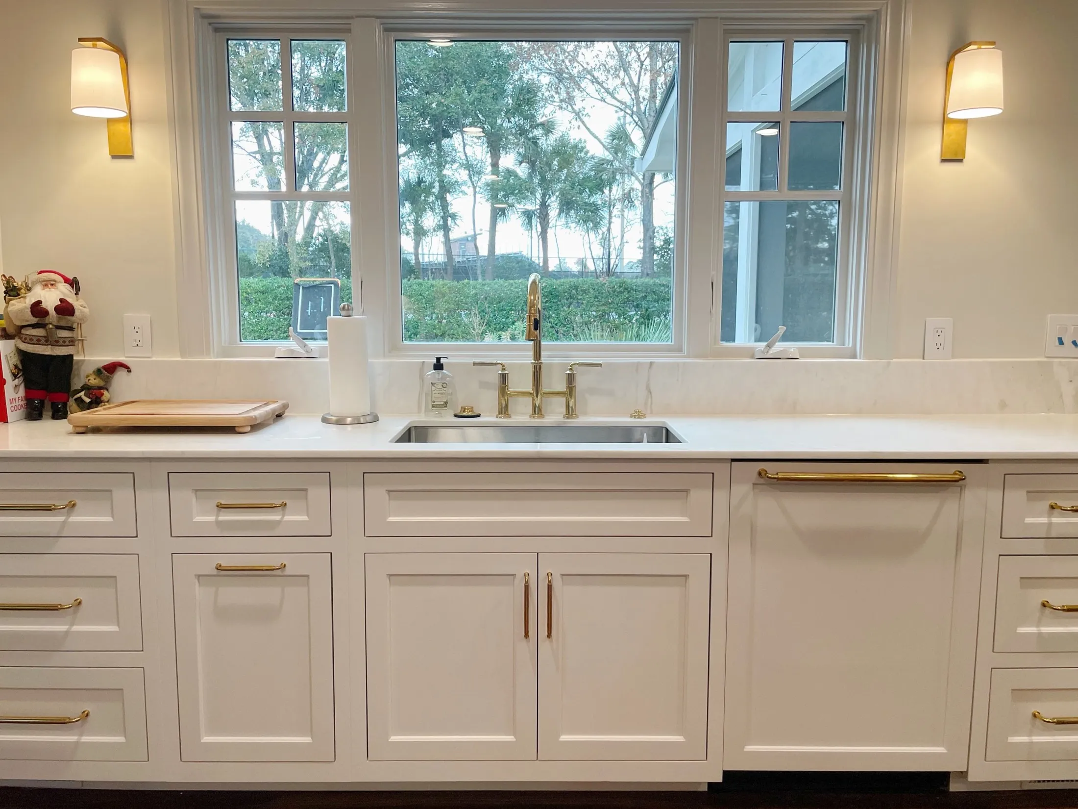 Kitchen sink with a window view of trees and outdoor scenery, decorated with Christmas figurines and a paper towel holder on the white countertop.