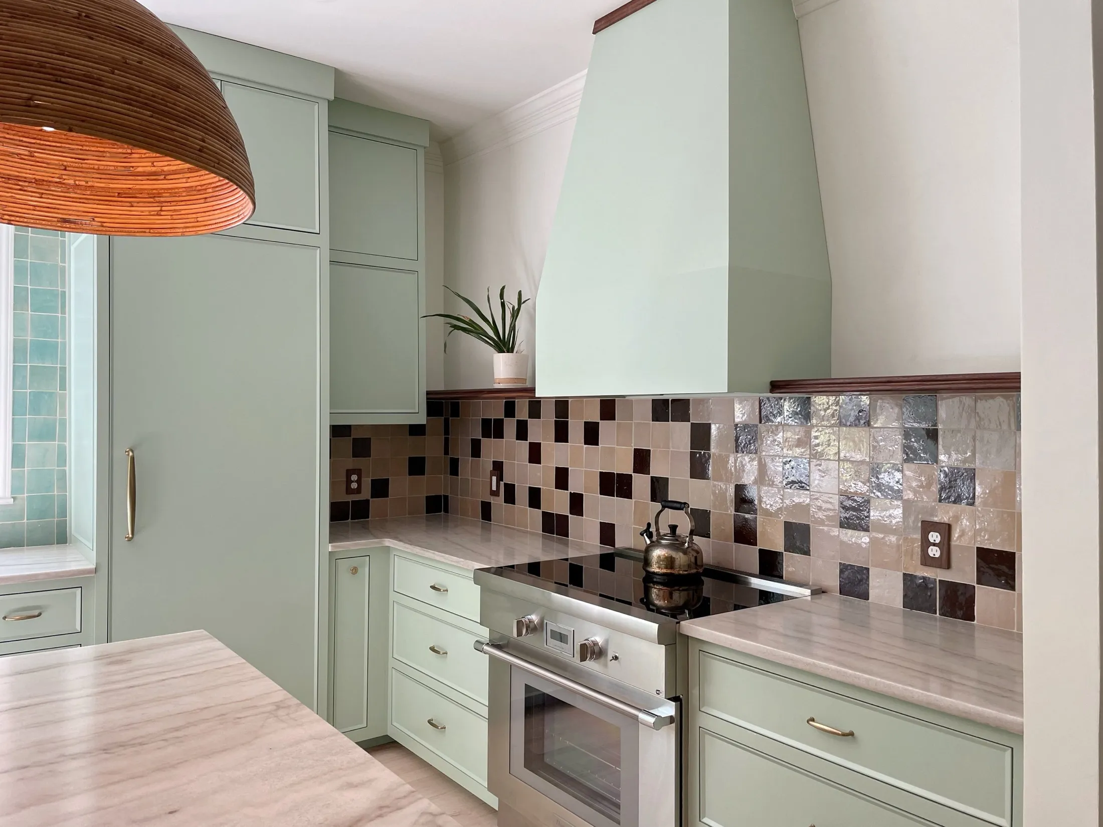 Kitchen with light green cabinets, a beige marble countertop, a multicolored tile backsplash, a built-in oven with a kettle on top, a potted plant on the corner, and a large wooden pendant light
