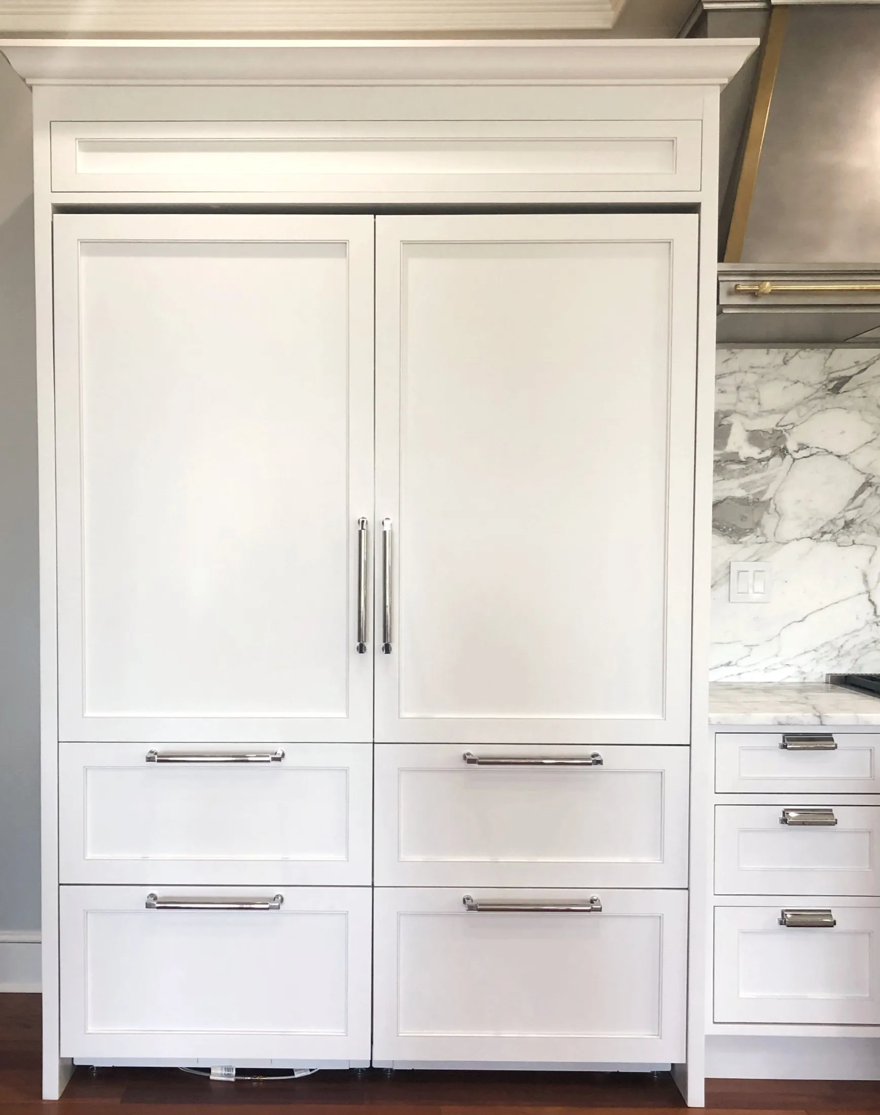 White kitchen cabinet with large central doors, three drawers below, and silver handles, next to marble backsplash and countertop.