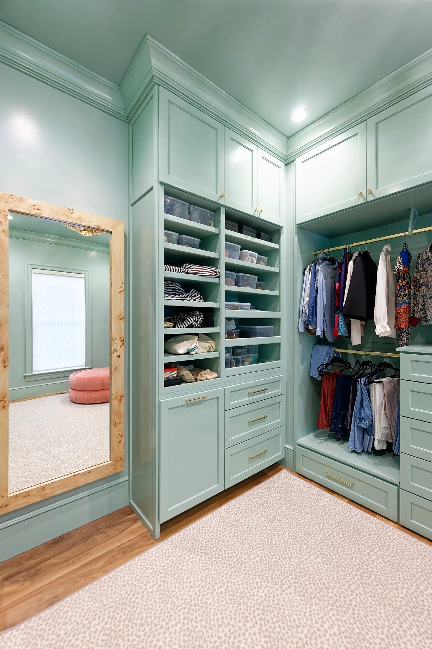 Walk-in closet with built-in shelves and hanging space filled with clothes. A full-length mirror reflects a pink ottoman in an adjoining room with beige carpet and green walls.