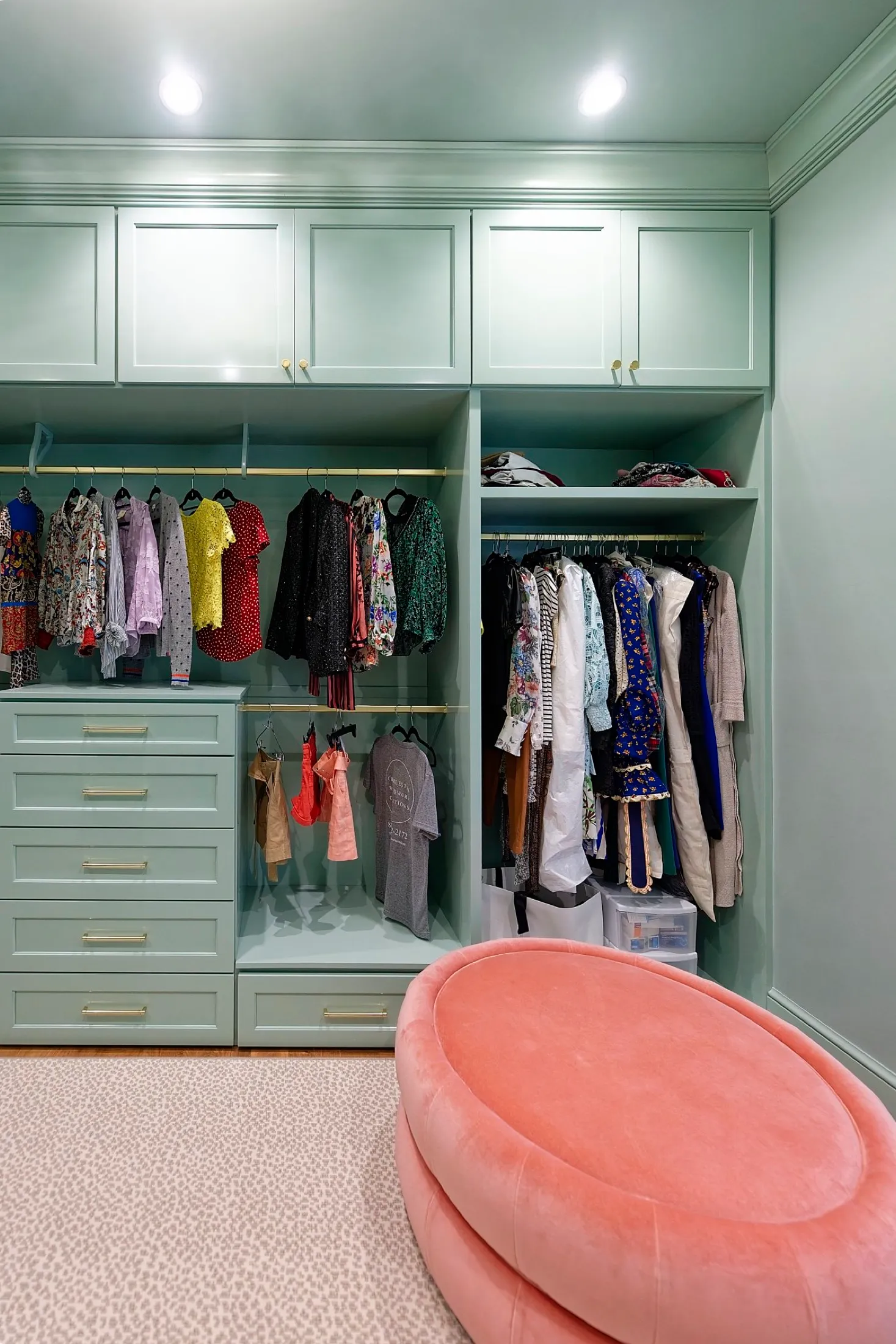 A walk-in closet with mint green cabinetry, hanging clothes, and a pink velvet bench in the foreground.