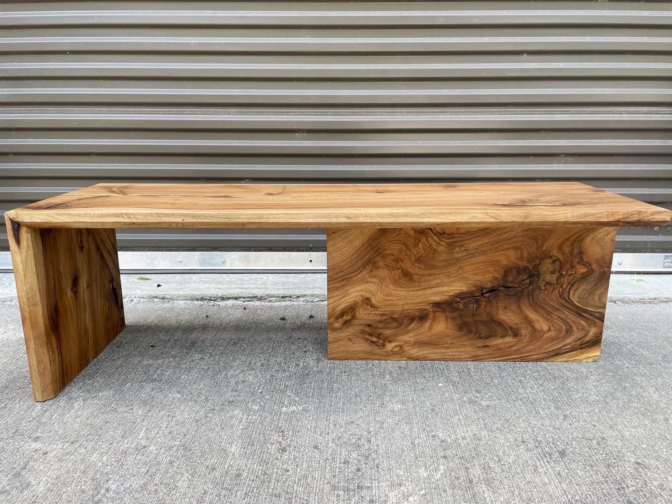 Wooden bench with a natural finish, featuring a rectangular seat and a large rectangular support on the right side, placed on a concrete surface in front of a metallic, corrugated wall.