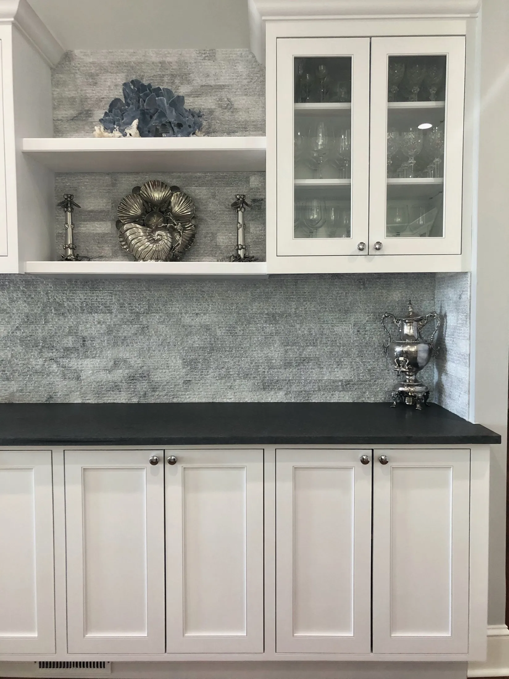 White kitchen cabinet with black countertop, glass-front upper cabinet with glassware, decorative silver teapot and silver decorative plates on shelves with gray textured backsplash.