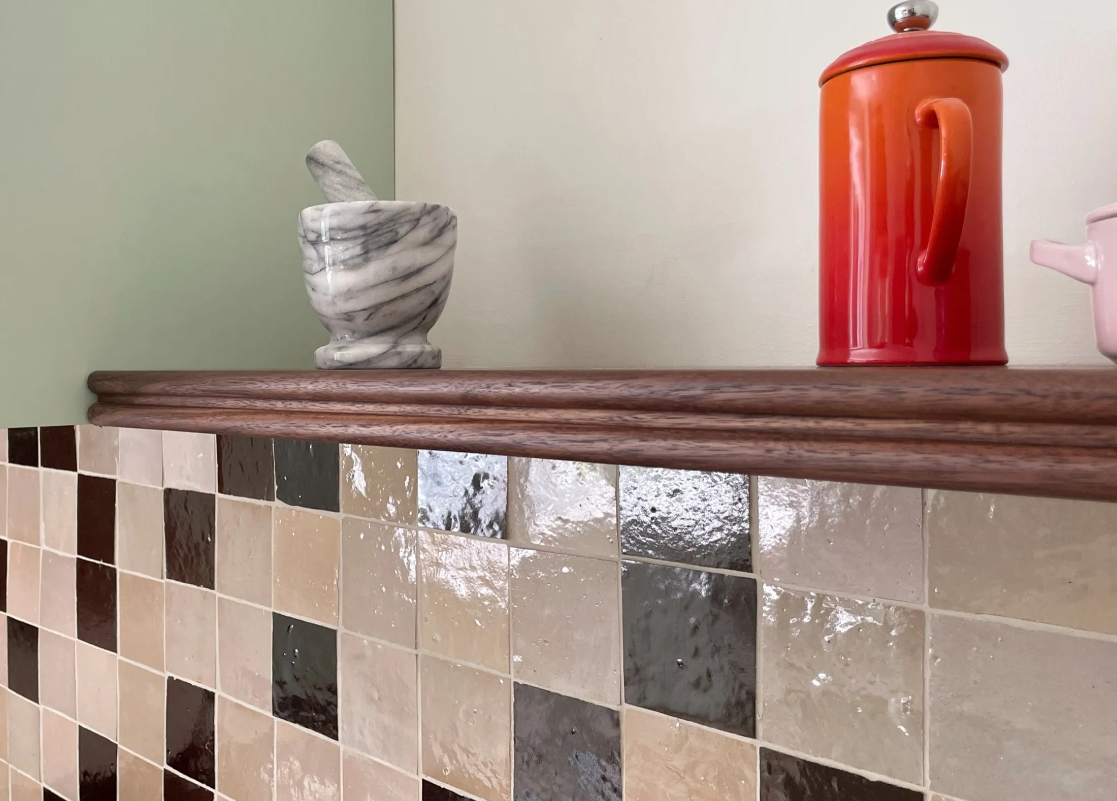 Decorative bathroom shelf with a marble mortar and pestle, an orange thermos, and a pink pitcher against a tiled wall.