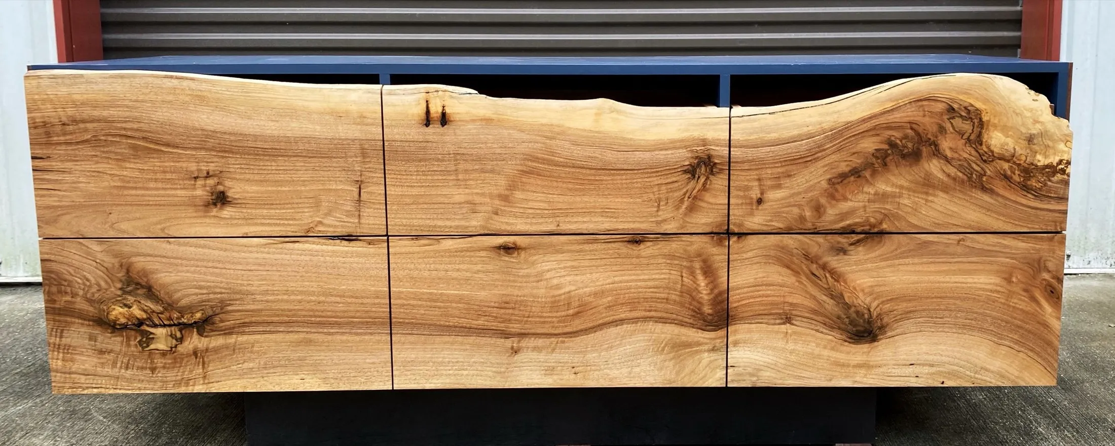 A wooden cabinet with a natural, live edge top, featuring six large, smooth, and polished wood panels with visible grain and knots, set outdoors on a concrete surface with a metallic fence and building background.