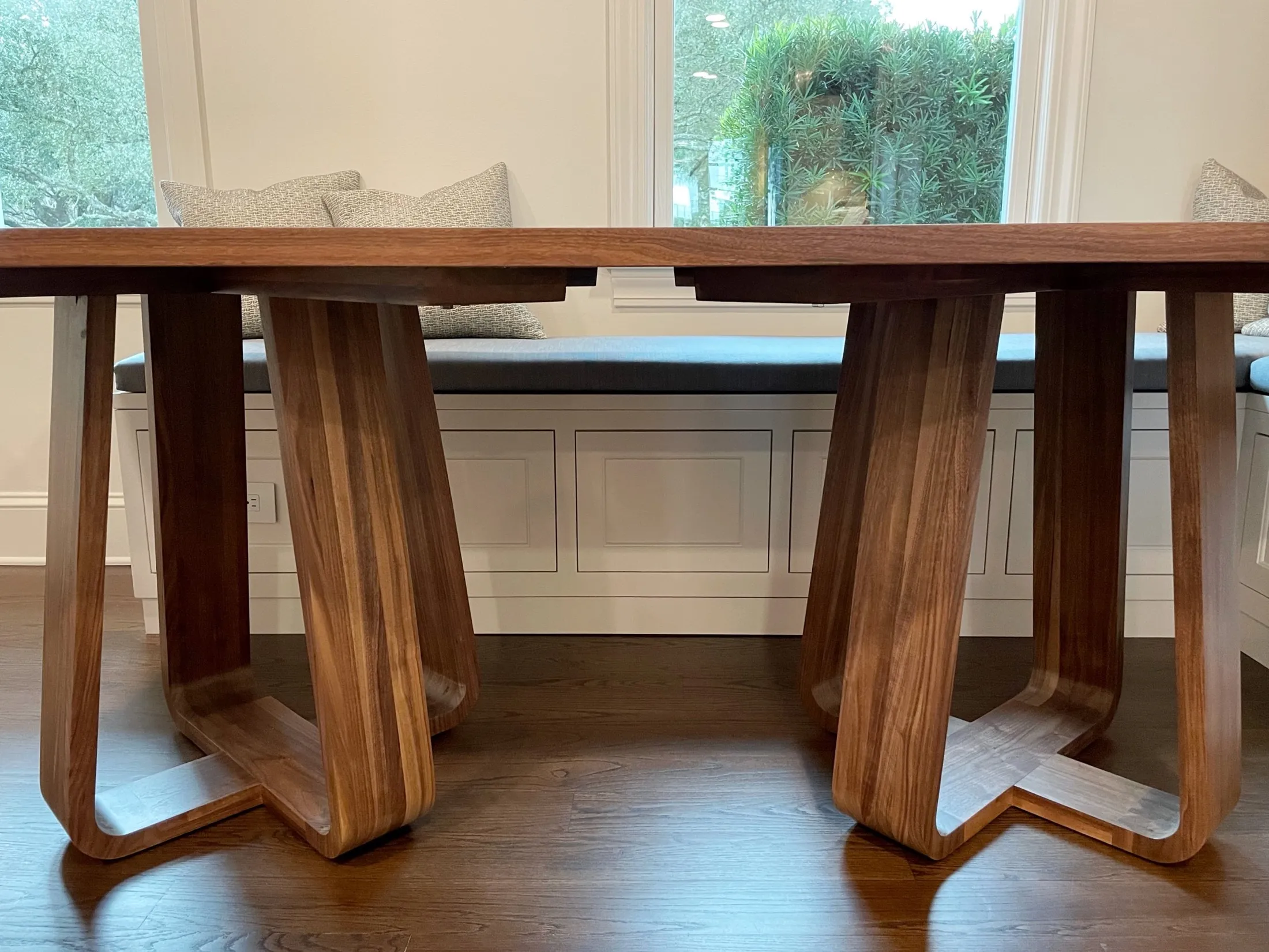 Wooden dining table with a modern design, positioned in a bright room with a window seat and large windows showing greenery outside.