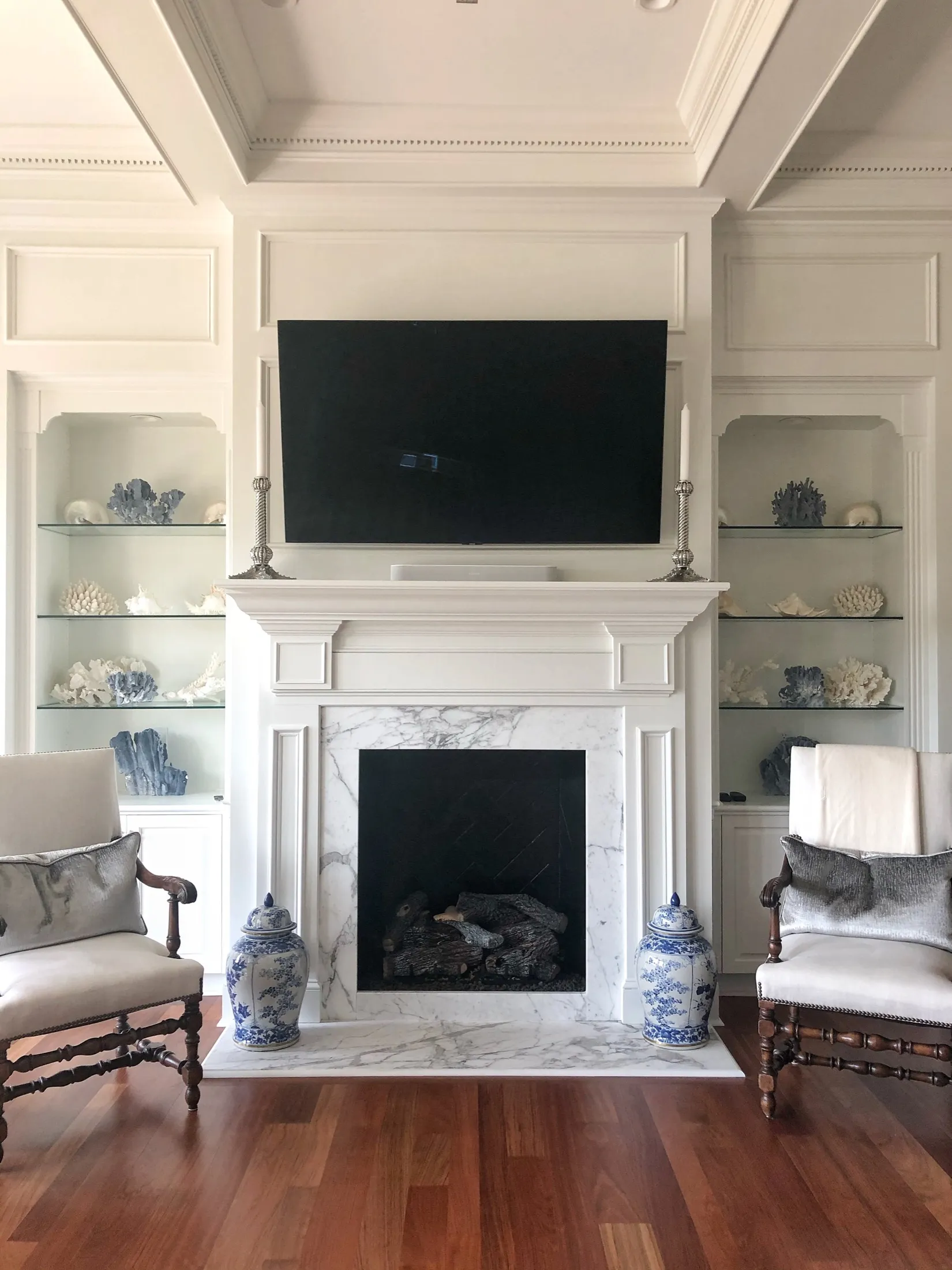 A living room with a built-in white wall unit, decorated with sea-themed ornaments in glass displays on either side. A flat-screen TV is mounted above a white marble fireplace with a black interior, flanked by two blue and white vases. Two antique-st