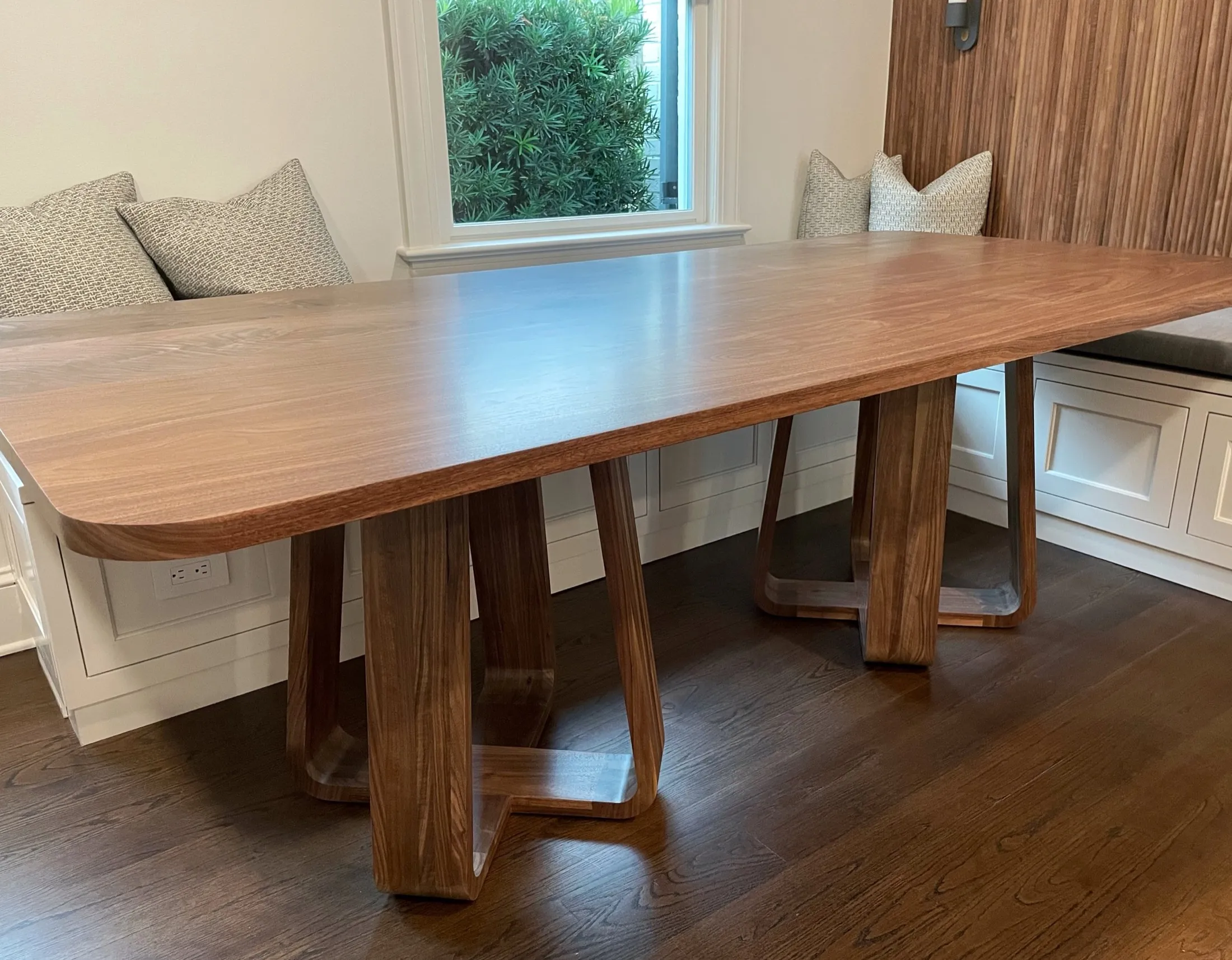 A modern wooden dining table with a thick, smooth tabletop and sculptural legs, positioned in a room with natural light from a window, with cushions on the built-in bench seating.
