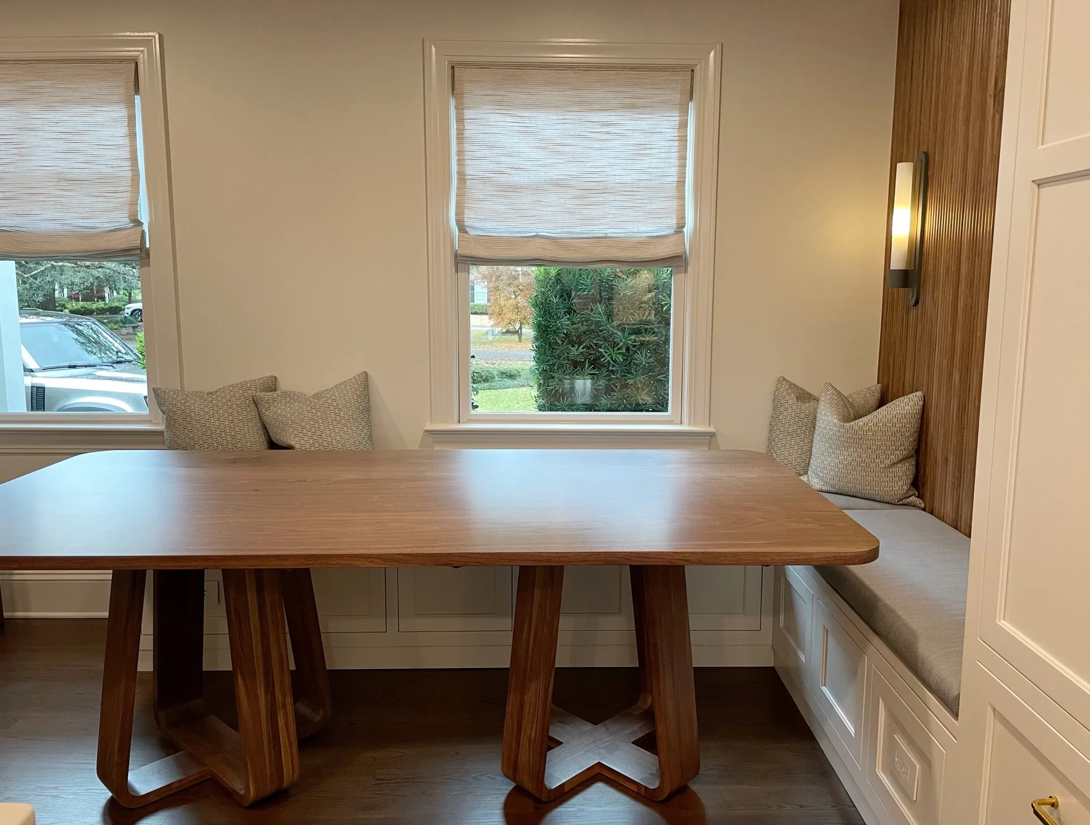 A woodworking nook with a wooden table and built-in bench with pillows, situated in front of two windows with Roman shades, and a wall sconce light.