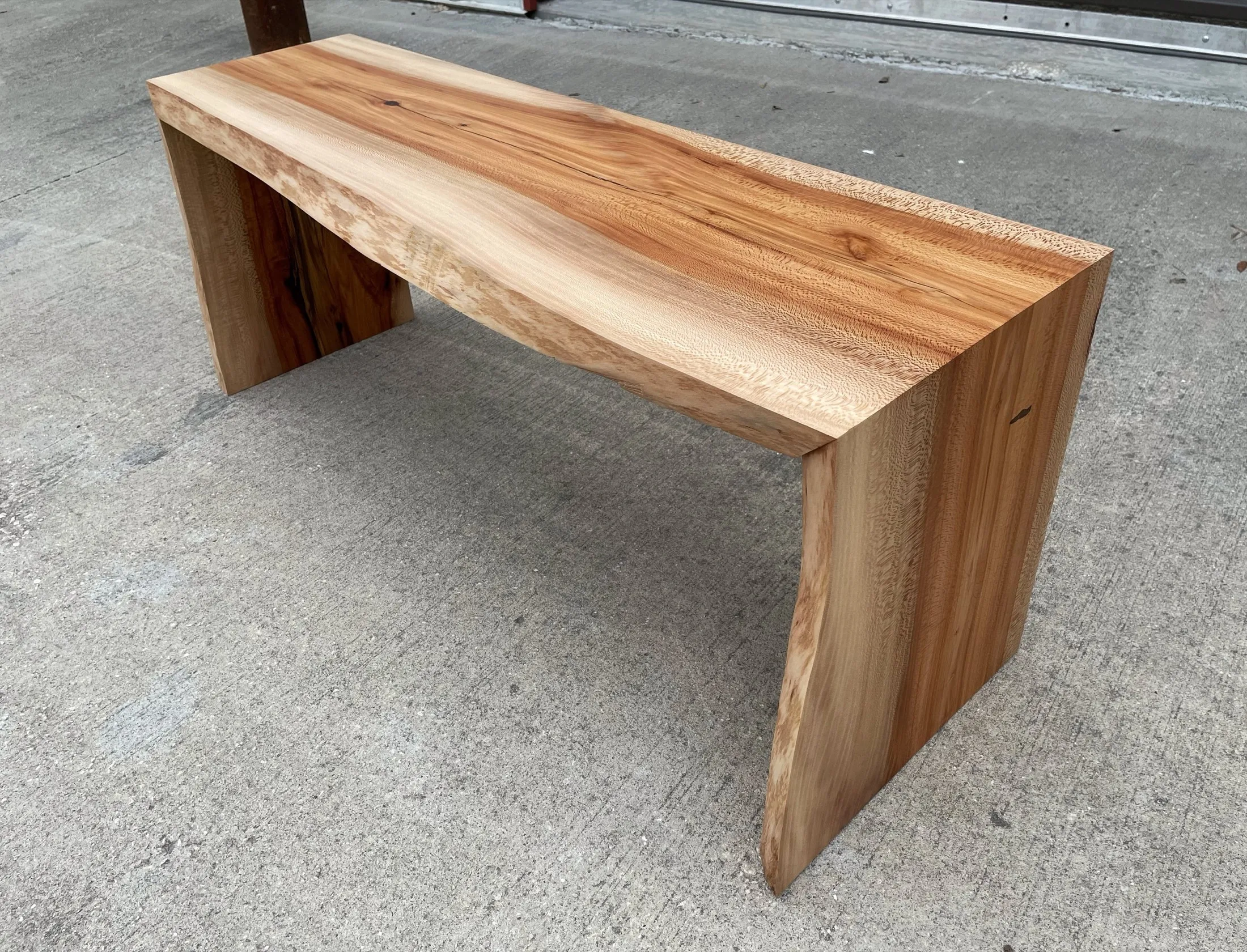 A wooden bench constructed from a single piece of natural wood with a smooth, polished finish, placed on a concrete sidewalk outside.