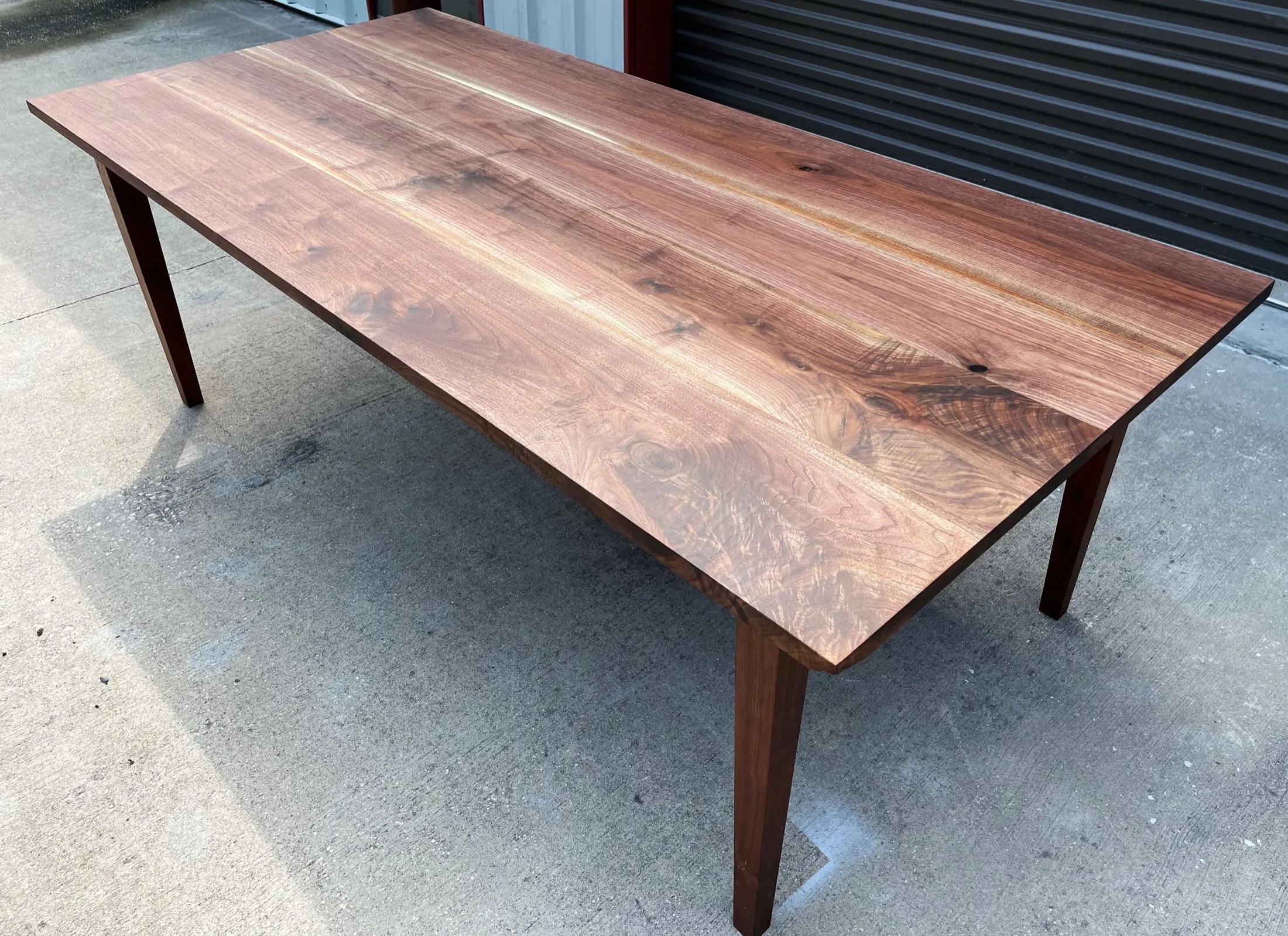 A wooden table with a polished, natural wood grain finish, situated outdoors on concrete pavement.