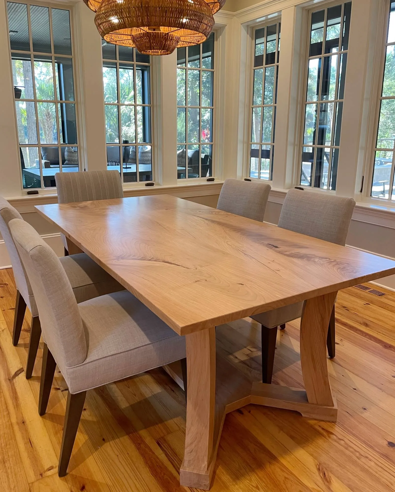American elm extension dining table with curved trestle legs, set with linen chairs in a sunny room lined with windows.