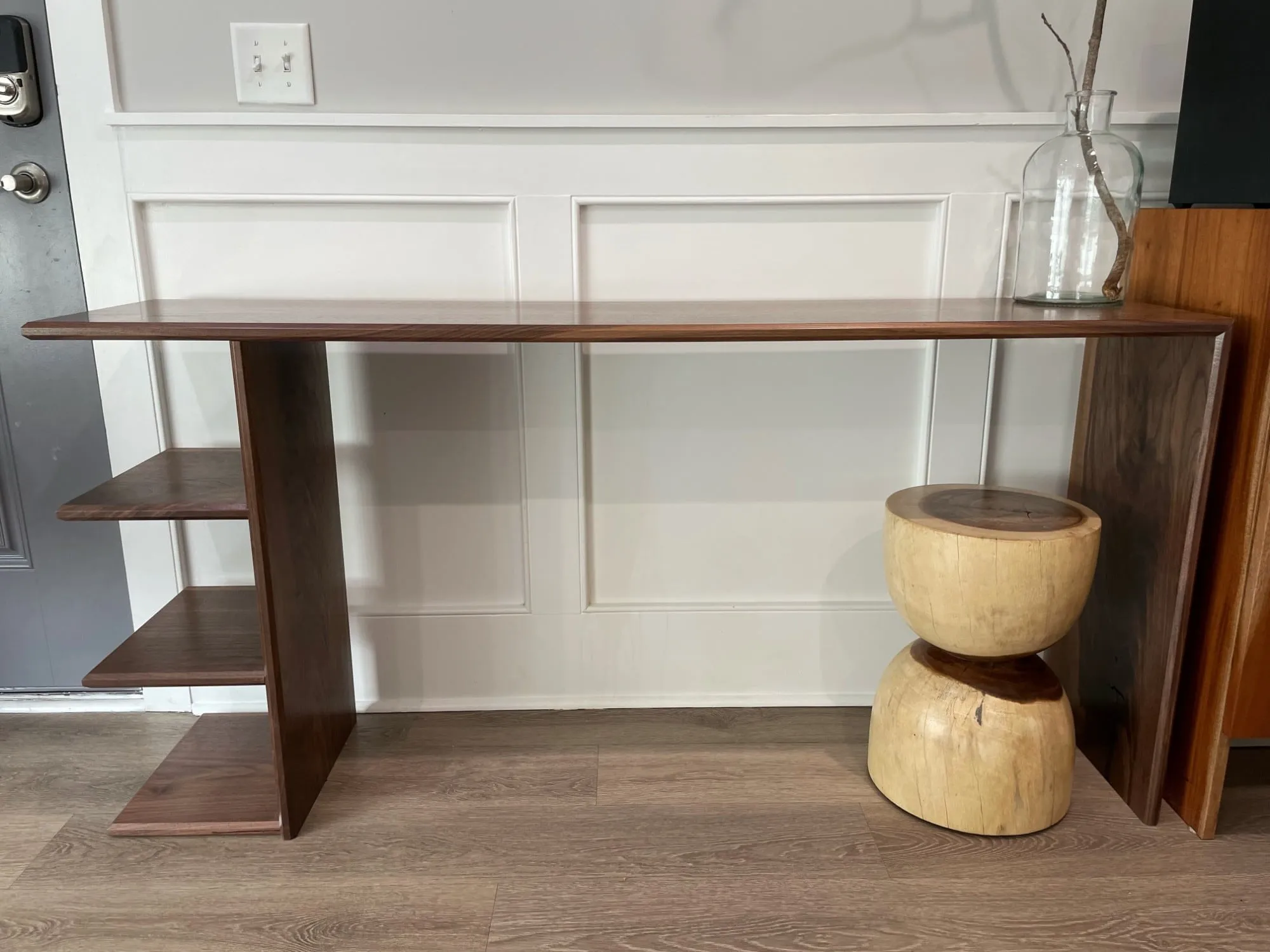 Walnut console table with a long top, open shelves stepping down one end, and a slab leg at the other, against a paneled wall.