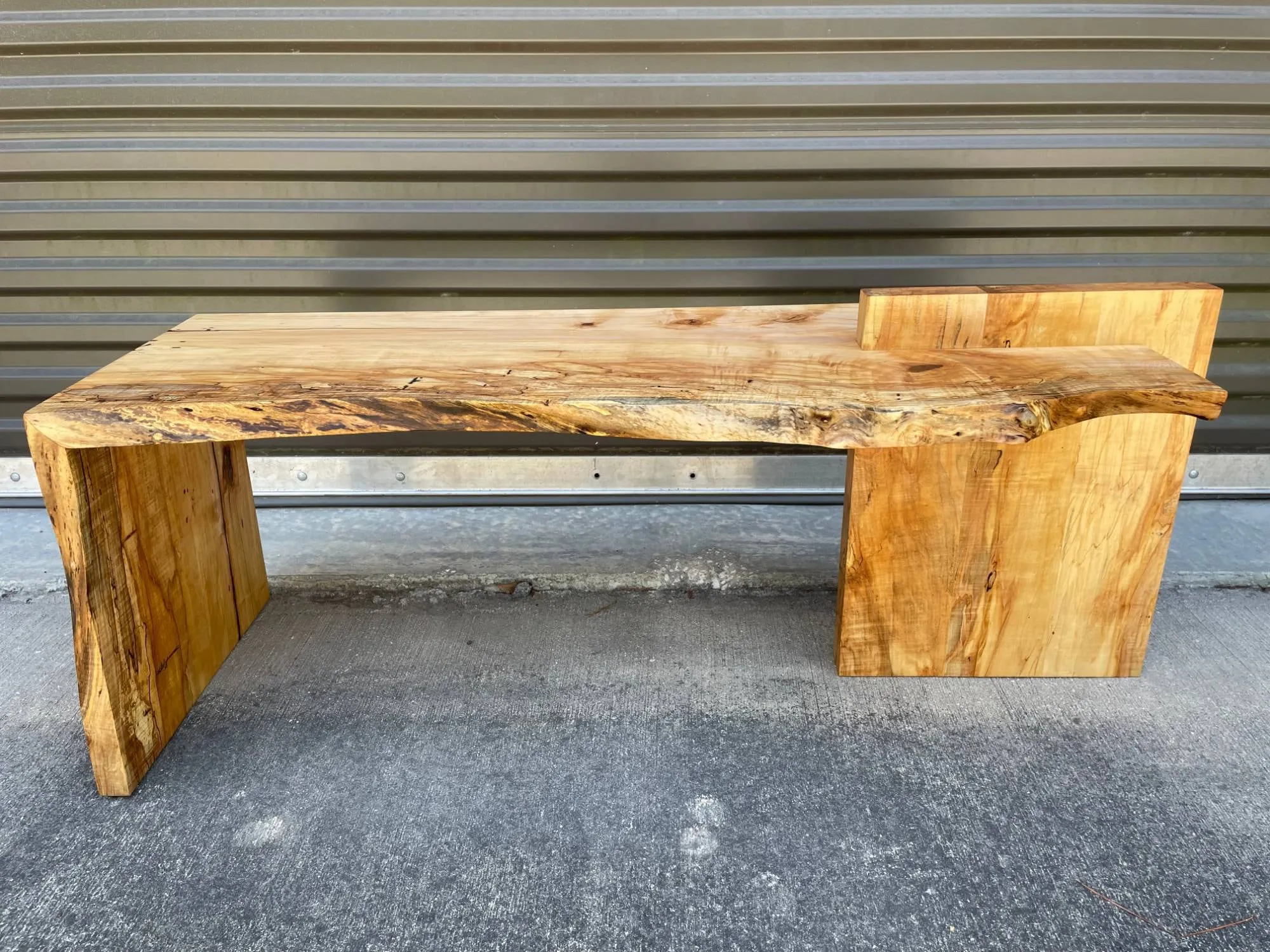 Spalted maple live-edge bench with a waterfall leg at one end and a block leg with a raised armrest at the other.