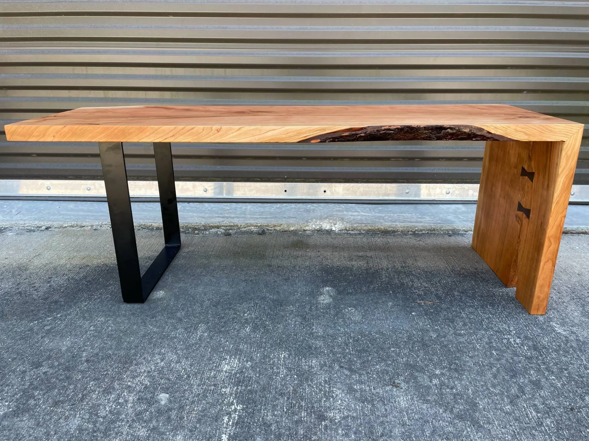Cherry live-edge bench with a black steel leg at one end and a waterfall leg with walnut bowtie inlays at the other.